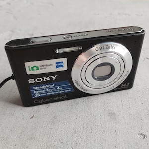 May include: A black Sony Cyber-shot digital camera featuring a silver Carl Zeiss Vario-Tessar lens, 14.1 megapixels, 4x optical zoom, and a 26mm wide-angle lens. The front displays Intelligent Auto branding and a SteadyShot label.