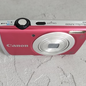 Op de afbeelding: Een roze en zilverkleurige Canon PowerShot A2500 digitale camera met een zilveren ontspanknop aan de bovenkant, een zwarte aan/uit-knop met het label ON/OFF en een lens. De camerabehuizing is voorzien van het Canon-logo en de modelnaam.