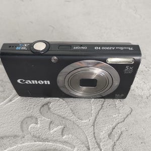 May include: A black Canon PowerShot A2300 HD digital camera featuring a 5x optical zoom lens, a 16.0 megapixel sensor, and a top-mounted silver shutter button. The camera body displays the brand name Canon and model text clearly.