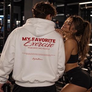 Puede incluir: Una sudadera con capucha blanca que presenta texto rojo en la parte posterior que dice MY FAVORITE Exercise REQUIRES TWO PEOPLE, junto con texto impreso más pequeño y un logotipo de firma. La prenda tiene capucha y mangas largas.