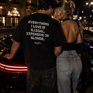 May include: A black t-shirt featuring the white text "EVERYTHING I LOVE IS ILLEGAL, EXPENSIVE OR BLONDE." and a signature logo. The shirt is paired with dark denim jeans, while a black backless top and light blue denim jeans are also visible.