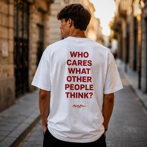 Puede incluir: Una camiseta blanca de manga corta con el texto rojo WHO CARES WHAT OTHER PEOPLE THINK? impreso en la espalda. Debajo del texto hay un logotipo más pequeño en letra cursiva roja que dice Nojta Mrae EST 2022. La camiseta tiene cuello redondo.