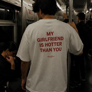 May include: A white short-sleeved t-shirt featuring the red text MY GIRLFRIEND IS HOTTER THAN YOU printed on the back. Below the text is a small red script logo. The shirt has a crew neckline and a relaxed fit, shown from the back view.
