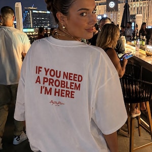 Könnte beinhalten: Ein weißes kurzärmeliges T-Shirt mit dem roten Text "IF YOU NEED A PROBLEM I'M HERE" auf der Rückseite, darunter befindet sich ein kleineres Logo in Schreibschrift. Das T-Shirt hat eine lockere Passform und wird mit einer blauen Jeans kombiniert.