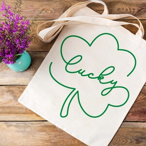 May include: A white canvas tote bag with a green outline of a four-leaf clover and the word "lucky" written in cursive.