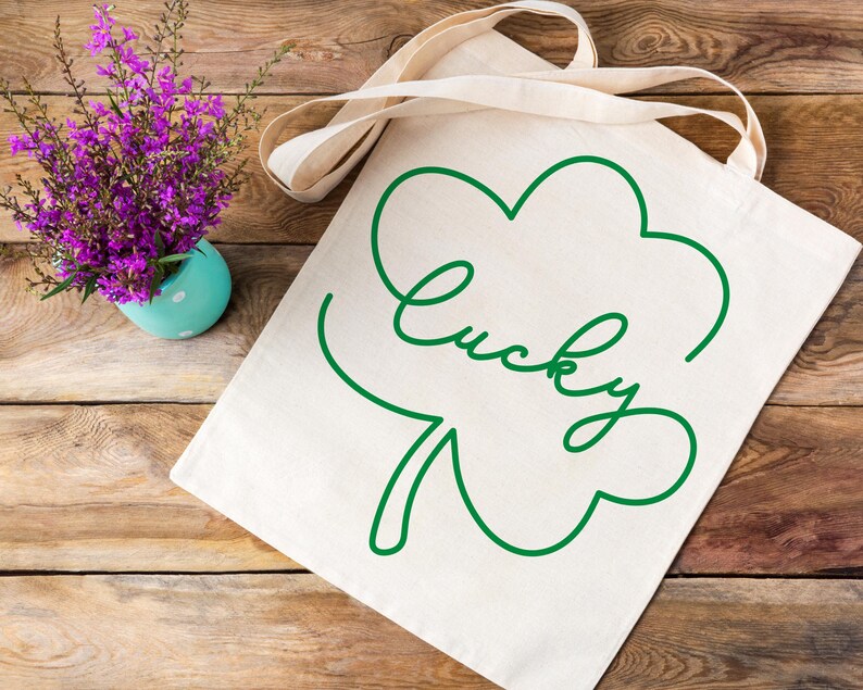 May include: A white canvas tote bag with a green outline of a four-leaf clover and the word "lucky" written in cursive.