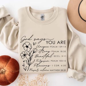 K&ouml;nnte beinhalten: Ein beiger Pullover mit schwarzem Text, der "Gott sagt, du bist Einzigartig, Stark, Sch&ouml;n, Vergeben, Kostbar, Niemals allein" lautet, mit Bibelversen, die unter jedem Wort aufgef&uuml;hrt sind. Der Text ist von einem floralen Design umgeben.