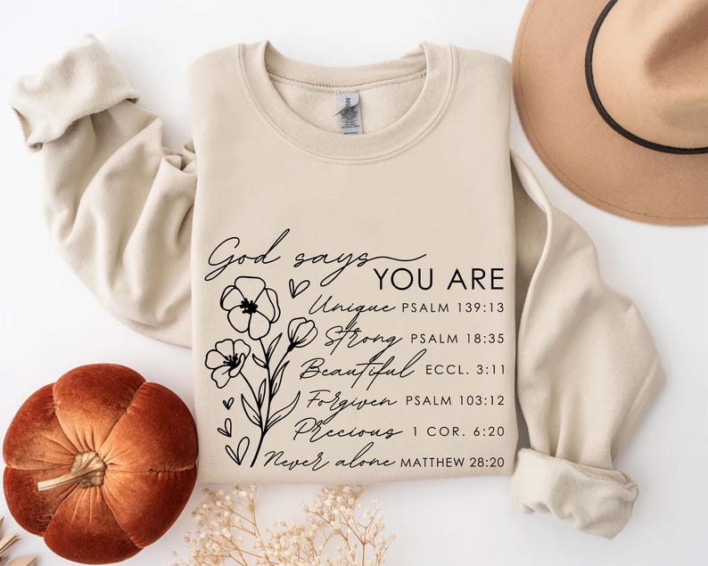 K&ouml;nnte beinhalten: Ein beiger Pullover mit schwarzem Text, der "Gott sagt, du bist Einzigartig, Stark, Sch&ouml;n, Vergeben, Kostbar, Niemals allein" lautet, mit Bibelversen, die unter jedem Wort aufgef&uuml;hrt sind. Der Text ist von einem floralen Design umgeben.