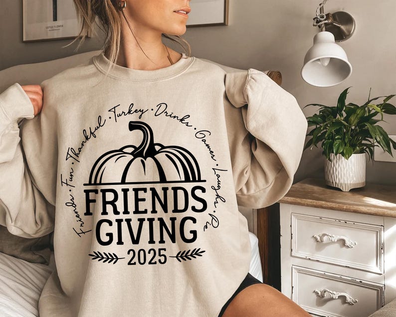 May include: Beige Friendsgiving sweatshirt featuring a black pumpkin graphic and the text "Friends Giving 2025". The design includes the words "Fun, Thankful, Turkey, Drinks, Games, Laughs, Pie" around the pumpkin.