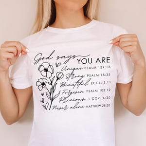 K&ouml;nnte beinhalten: Wei&szlig;es T-Shirt mit schwarzem Text und floralem Design. Der Text lautet "Gott sagt DU BIST Einzigartig Stark Sch&ouml;n Vergeben Wertvoll Niemals allein" mit Bibelversen, die unter jedem Wort aufgef&uuml;hrt sind.