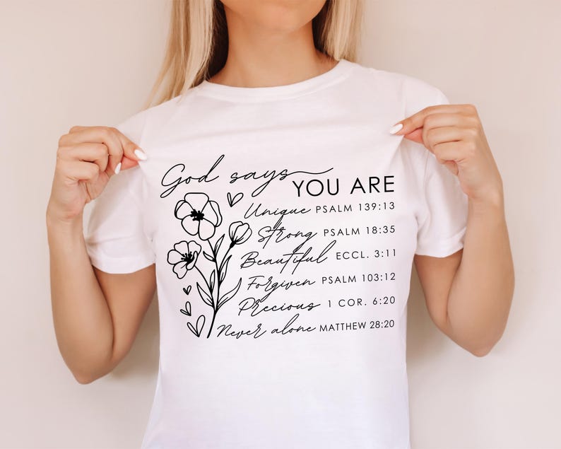 K&ouml;nnte beinhalten: Wei&szlig;es T-Shirt mit schwarzem Text und floralem Design. Der Text lautet "Gott sagt DU BIST Einzigartig Stark Sch&ouml;n Vergeben Wertvoll Niemals allein" mit Bibelversen, die unter jedem Wort aufgef&uuml;hrt sind.