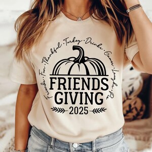 May include: A cream-coloured t-shirt with a black graphic. The design includes a pumpkin above the words "FRIENDS GIVING" and the year "2025". The words "Friends, Fun, Thankful, Turkey, Drinks, Games, Laughs, Pie" are in a circle.