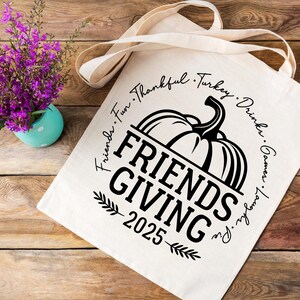 May include: A cream tote bag featuring a pumpkin graphic and the text "FRIENDS GIVING 2025". The words "Friends, Fun, Thankful, Turkey, Drinks, Games, Laughs, Pie" are printed around the pumpkin. A small turquoise vase with purple flowers is on the left.