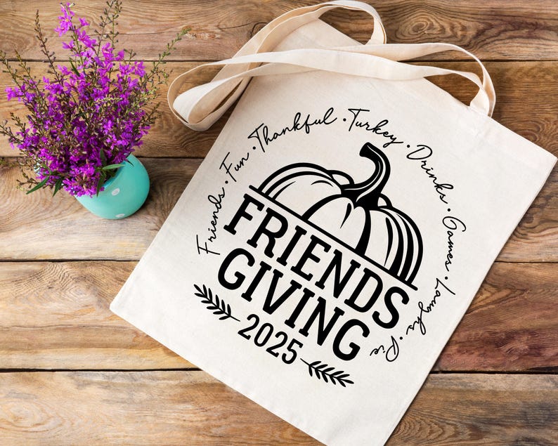 May include: A cream tote bag featuring a pumpkin graphic and the text "FRIENDS GIVING 2025". The words "Friends, Fun, Thankful, Turkey, Drinks, Games, Laughs, Pie" are printed around the pumpkin. A small turquoise vase with purple flowers is on the left.