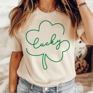 May include: A beige t-shirt with a green outline of a four-leaf clover and the word "lucky" written inside the clover.