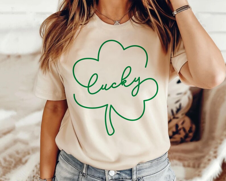 May include: A beige t-shirt with a green outline of a four-leaf clover and the word "lucky" written inside the clover.