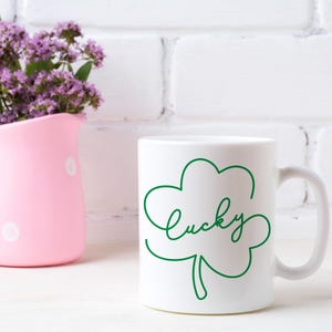 May include: White ceramic mug with a green shamrock design and the word "lucky" written in cursive. The mug is sitting on a white surface.