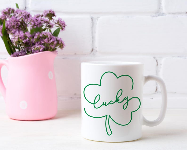 May include: White ceramic mug with a green shamrock design and the word "lucky" written in cursive. The mug is sitting on a white surface.