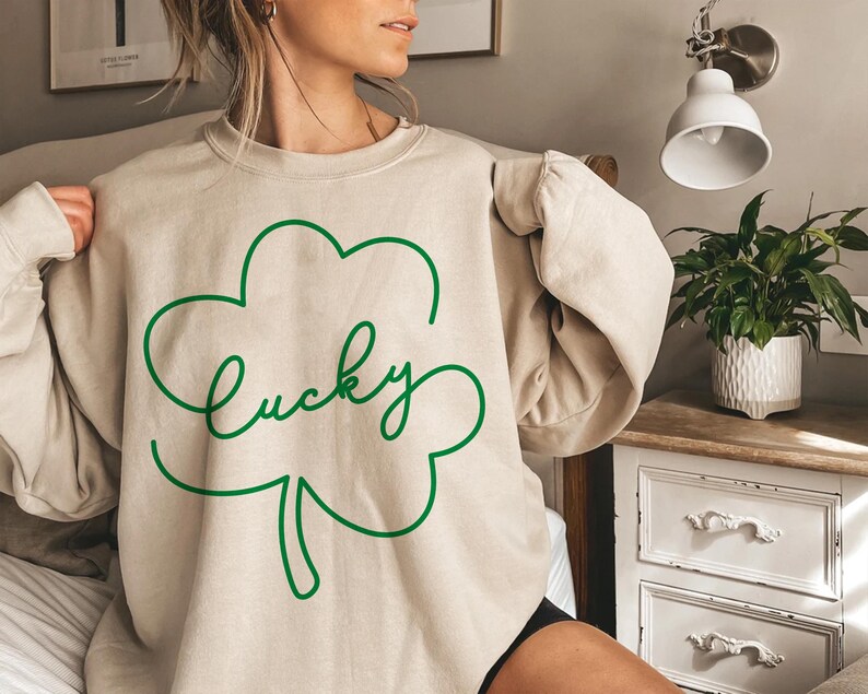 May include: A beige sweatshirt with a green outline of a four-leaf clover and the word "lucky" written inside the clover.