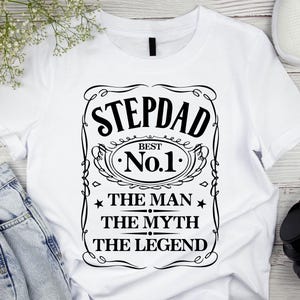 Pode incluir: Camiseta branca com um gr&aacute;fico preto que diz "STEPDAD BEST No.1 THE MAN THE MYTH THE LEGEND". O design &eacute; emoldurado com linhas decorativas e estrelas. A camiseta &eacute; exibida com jeans azuis, t&ecirc;nis brancos e fones de ouvido pretos.