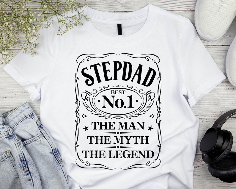 Pode incluir: Camiseta branca com um gr&aacute;fico preto que diz "STEPDAD BEST No.1 THE MAN THE MYTH THE LEGEND". O design &eacute; emoldurado com linhas decorativas e estrelas. A camiseta &eacute; exibida com jeans azuis, t&ecirc;nis brancos e fones de ouvido pretos.