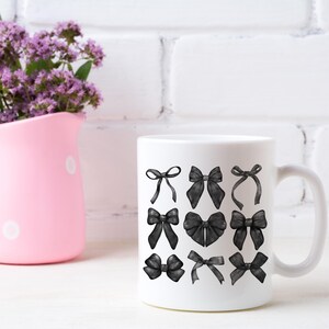 May include: White ceramic mug with a black and white illustration of various bows. The mug is sitting on a white surface with a pink pitcher and purple flowers in the background.