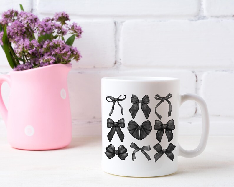 May include: White ceramic mug with a black and white illustration of various bows. The mug is sitting on a white surface with a pink pitcher and purple flowers in the background.