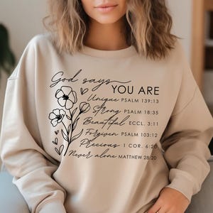 K&ouml;nnte beinhalten: Ein beiger Pullover mit schwarzem Text, der "Gott sagt DU BIST Einzigartig, Stark, Sch&ouml;n, Vergeben, Kostbar, Niemals allein" lautet, mit Bibelversen, die unter jedem Wort aufgef&uuml;hrt sind.