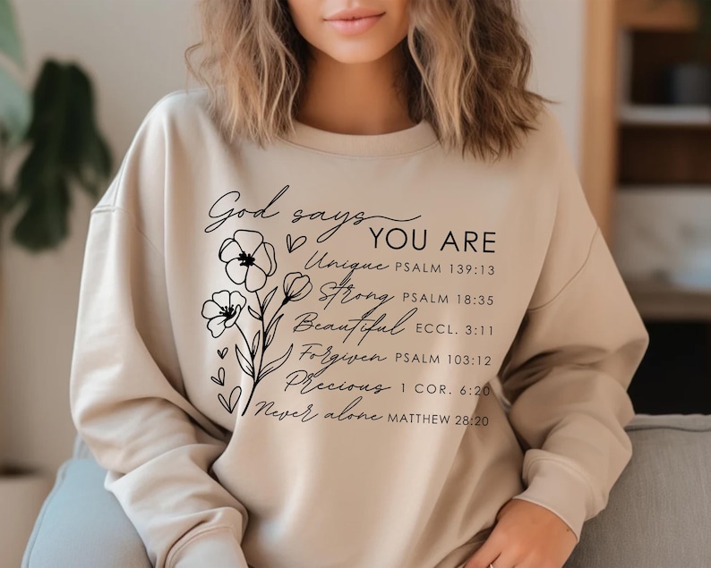 K&ouml;nnte beinhalten: Ein beiger Pullover mit schwarzem Text, der "Gott sagt DU BIST Einzigartig, Stark, Sch&ouml;n, Vergeben, Kostbar, Niemals allein" lautet, mit Bibelversen, die unter jedem Wort aufgef&uuml;hrt sind.