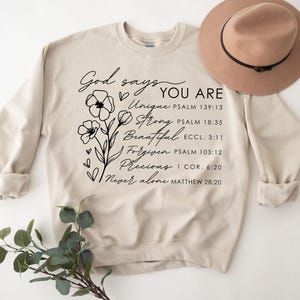 K&ouml;nnte beinhalten: Ein beiger Pullover mit einem schwarzen Blumenmuster und dem Text "Gott sagt, du bist Einzigartig, Stark, Sch&ouml;n, Vergeben, Kostbar, Niemals allein" mit Bibelversen, die unter jedem Wort aufgef&uuml;hrt sind.