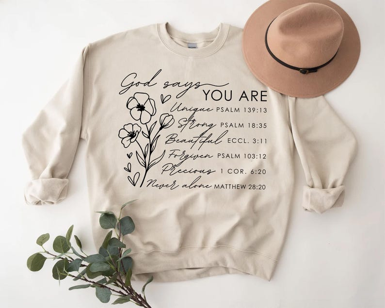 K&ouml;nnte beinhalten: Ein beiger Pullover mit einem schwarzen Blumenmuster und dem Text "Gott sagt, du bist Einzigartig, Stark, Sch&ouml;n, Vergeben, Kostbar, Niemals allein" mit Bibelversen, die unter jedem Wort aufgef&uuml;hrt sind.