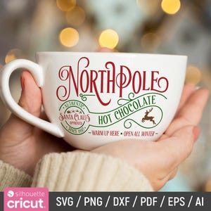 May include: White ceramic mug with a red and green design that says "North Pole Hot Chocolate" with a reindeer illustration. The text also says "Authentic Santa Claus Approved Since 1897" and "Warm Up Here Open All Winter".
