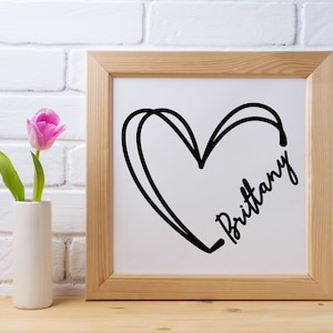 May include: A framed print with a black and white illustration of a heart with the name "Brittany" written inside. The frame is light brown wood and the background is a white brick wall.