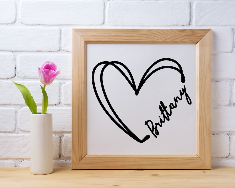 May include: A framed print with a black and white illustration of a heart with the name "Brittany" written inside. The frame is light brown wood and the background is a white brick wall.