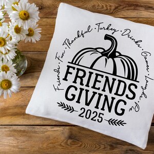 May include: A white cushion featuring a black pumpkin design and the text "Friends Giving 2025". The words "Friends, Fun, Thankful, Turkey, Drinks, Games, Laugh, Pie" are written around the pumpkin. Daisies in a glass vase are in the background.