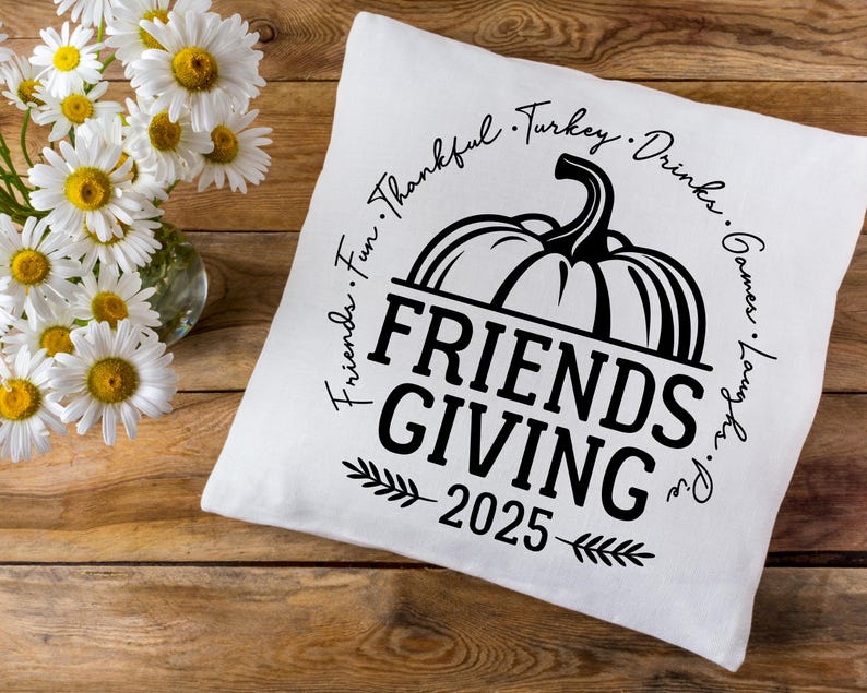 May include: A white cushion featuring a black pumpkin design and the text "Friends Giving 2025". The words "Friends, Fun, Thankful, Turkey, Drinks, Games, Laugh, Pie" are written around the pumpkin. Daisies in a glass vase are in the background.