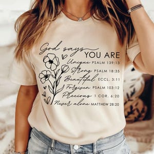 K&ouml;nnte beinhalten: Ein beiges T-Shirt mit schwarzem Text, der lautet: "God says YOU ARE Unique PSALM 139:13 Strong PSALM 18:35 Beautiful ECCL. 3:11 Forgiven PSALM 103:12 Precious I COR. 6:20 Never alone MATTHEW 28:20". Der Text ist von einem floralen Design umgeben.