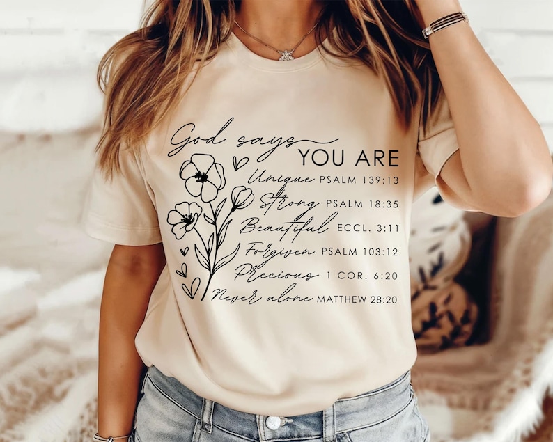 K&ouml;nnte beinhalten: Ein beiges T-Shirt mit schwarzem Text, der lautet: "God says YOU ARE Unique PSALM 139:13 Strong PSALM 18:35 Beautiful ECCL. 3:11 Forgiven PSALM 103:12 Precious I COR. 6:20 Never alone MATTHEW 28:20". Der Text ist von einem floralen Design umgeben.