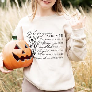 K&ouml;nnte beinhalten: Ein cremefarbener Pullover mit dem Text "God says YOU ARE" und einer Liste positiver Affirmationen mit entsprechenden Bibelversen, die darauf gedruckt sind. Der Text ist in schwarzer kursivschrift und enth&auml;lt die W&ouml;rter "Unique", "Strong", "Beautiful", "Forgiven", "Precious" und "Never alone".