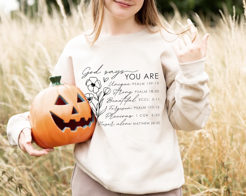 K&ouml;nnte beinhalten: Ein cremefarbener Pullover mit dem Text "God says YOU ARE" und einer Liste positiver Affirmationen mit entsprechenden Bibelversen, die darauf gedruckt sind. Der Text ist in schwarzer kursivschrift und enth&auml;lt die W&ouml;rter "Unique", "Strong", "Beautiful", "Forgiven", "Precious" und "Never alone".