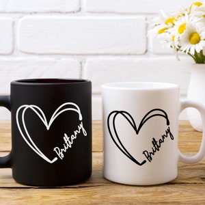 May include: Two ceramic coffee mugs with black and white designs. The mugs have a heart shape with the name "Brittany" written inside. One mug is black with white text and the other is white with black text.