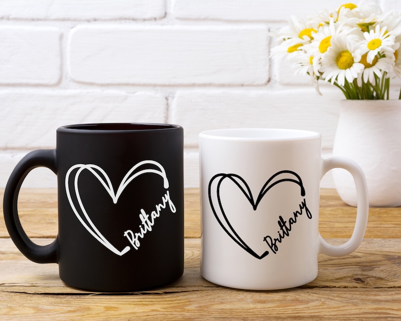 May include: Two ceramic coffee mugs with black and white designs. The mugs have a heart shape with the name "Brittany" written inside. One mug is black with white text and the other is white with black text.