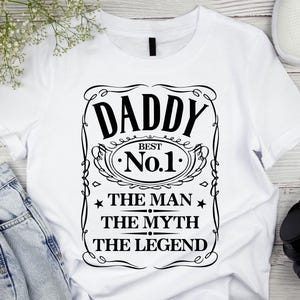 Pode incluir: T-shirt branca com um gr&aacute;fico preto que diz "DADDY BEST No.1 THE MAN THE MYTH THE LEGEND" em uma moldura decorativa. A camisa &eacute; feita de um material macio e &eacute; adequada para uso casual.