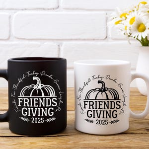 May include: Two ceramic mugs, one black and one white, with a Thanksgiving theme. Each mug features a pumpkin graphic and the words "FRIENDS GIVING 2025" in a circular design. A bouquet of daisies sits in a white vase.