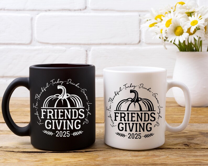 May include: Two ceramic mugs, one black and one white, with a Thanksgiving theme. Each mug features a pumpkin graphic and the words "FRIENDS GIVING 2025" in a circular design. A bouquet of daisies sits in a white vase.
