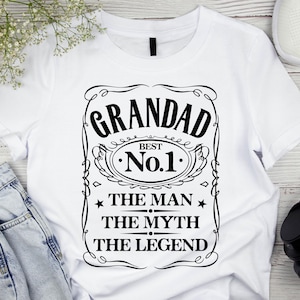 Pode incluir: Camiseta branca com um gr&aacute;fico preto que diz "GRANDAD BEST No.1 THE MAN THE MYTH THE LEGEND". O design &eacute; emoldurado com linhas decorativas e estrelas. A camiseta &eacute; exibida com jeans, fones de ouvido e t&ecirc;nis.