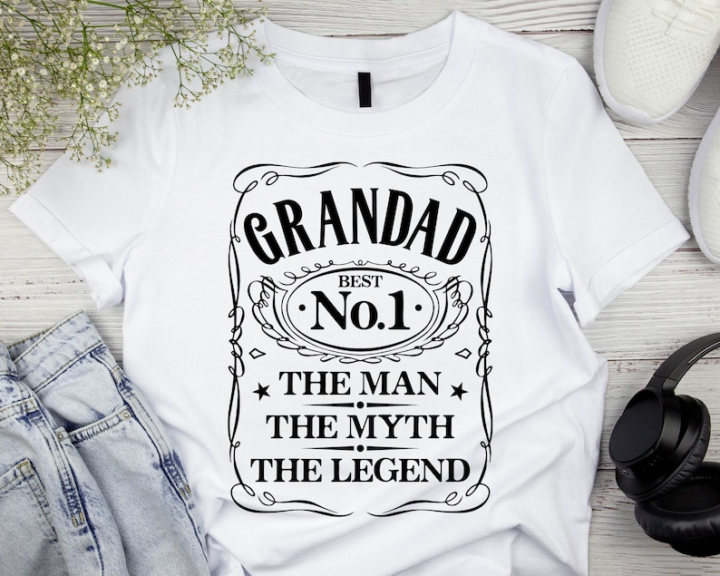 Pode incluir: Camiseta branca com um gr&aacute;fico preto que diz "GRANDAD BEST No.1 THE MAN THE MYTH THE LEGEND". O design &eacute; emoldurado com linhas decorativas e estrelas. A camiseta &eacute; exibida com jeans, fones de ouvido e t&ecirc;nis.