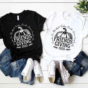May include: Two t-shirts, one black and one white, featuring the text "Friends Giving 2025" and a pumpkin design. The shirts are styled with blue denim jeans and white trainers, on a white wooden surface.