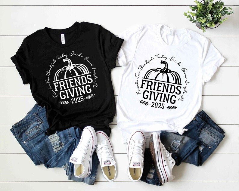 May include: Two t-shirts, one black and one white, featuring the text "Friends Giving 2025" and a pumpkin design. The shirts are styled with blue denim jeans and white trainers, on a white wooden surface.