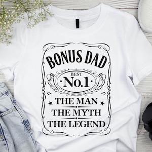 Pode incluir: Camiseta branca com um gr&aacute;fico preto que diz "BONUS DAD BEST No.1 THE MAN THE MYTH THE LEGEND". O design &eacute; emoldurado com linhas decorativas e estrelas. A camiseta &eacute; exibida em uma superf&iacute;cie branca com um par de jeans azuis, t&ecirc;nis brancos e fones de ouvido pretos.